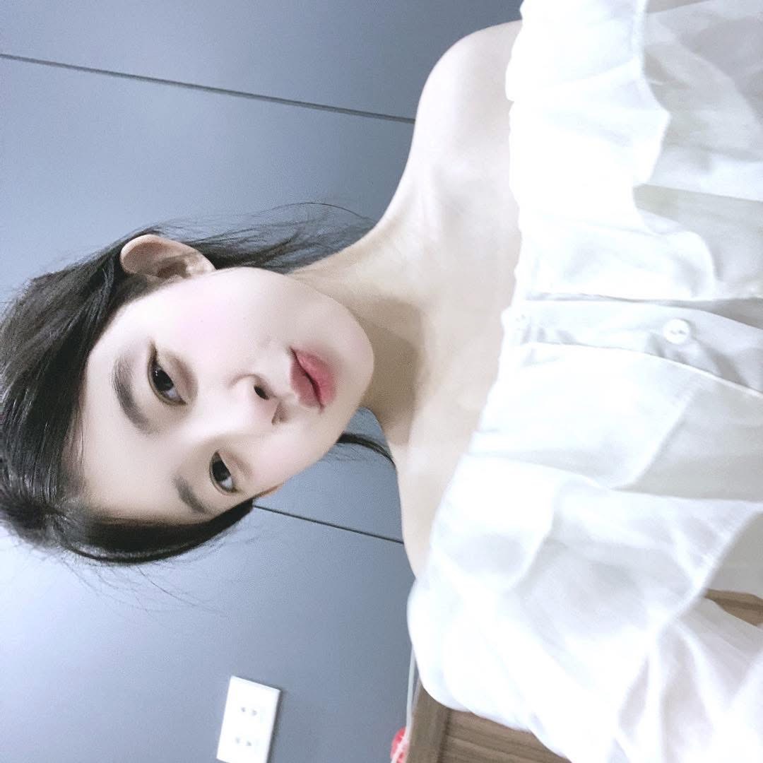 A young girl with long black hair wearing a white off-shoulder top, posing for a selfie with her head tilted, looking directly at the camera and making a cute pout.